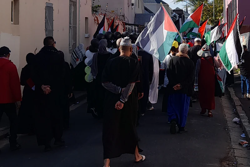 A Palestine solidarity march in Cape Town in June 2025, seen from behind, with Palestinian flags carried up a narrow street.
