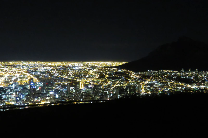 The lights of the Cape Town city bowl at night, seen from Signal Hill, with Table Mountain dark behind them.